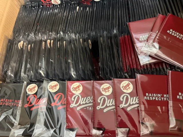 Rows of packaged red and black Duesy's Tavern koozies with logo prints and round branding stickers
